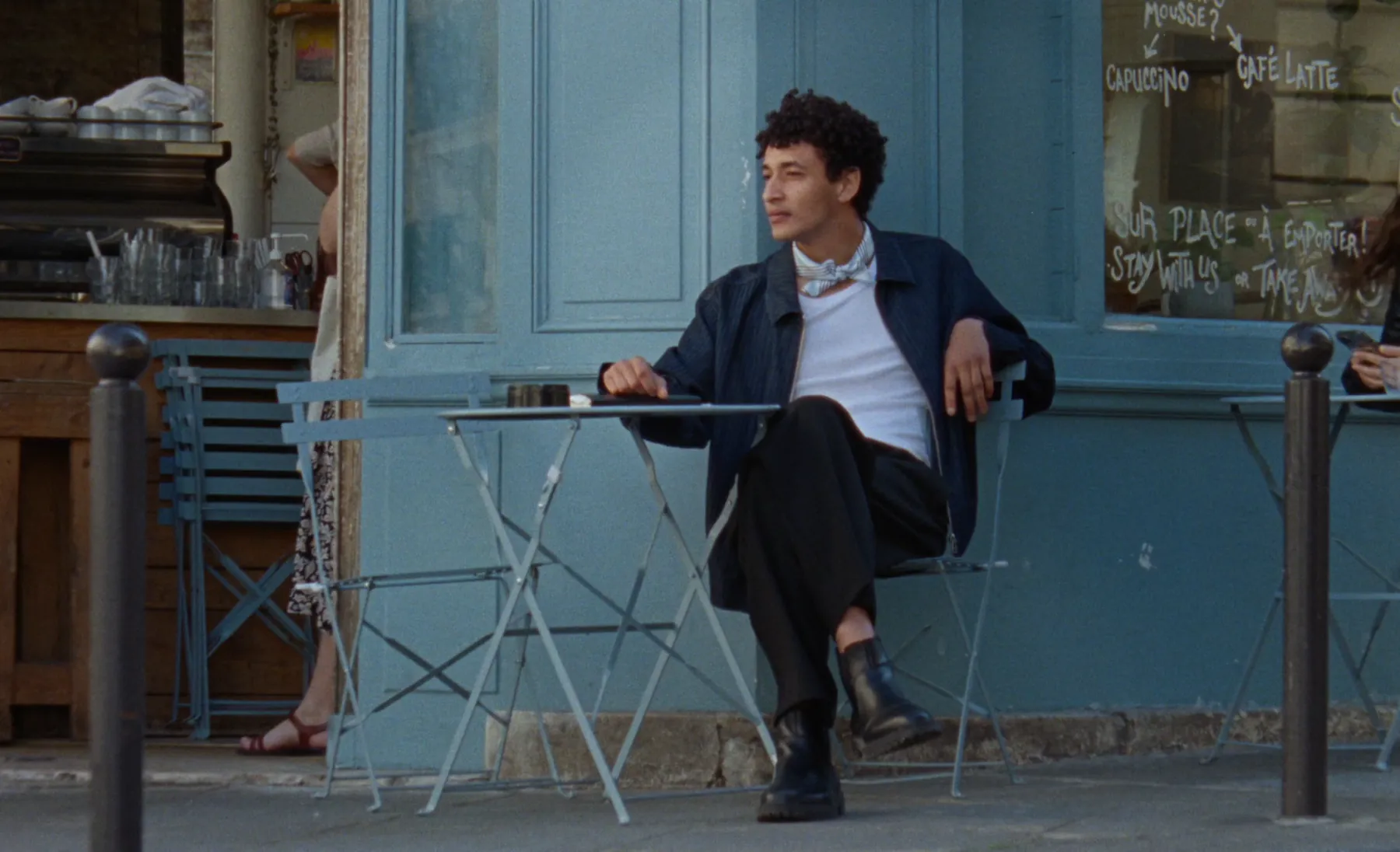A man seated at a café table, looking off toward the street